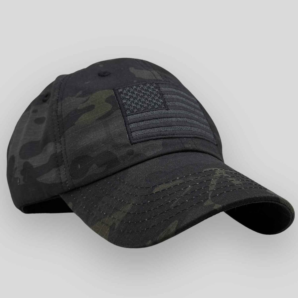 A black multicam camouflage hat cap featuring an embroidered black American flag patch on the front, set against a plain light gray background.