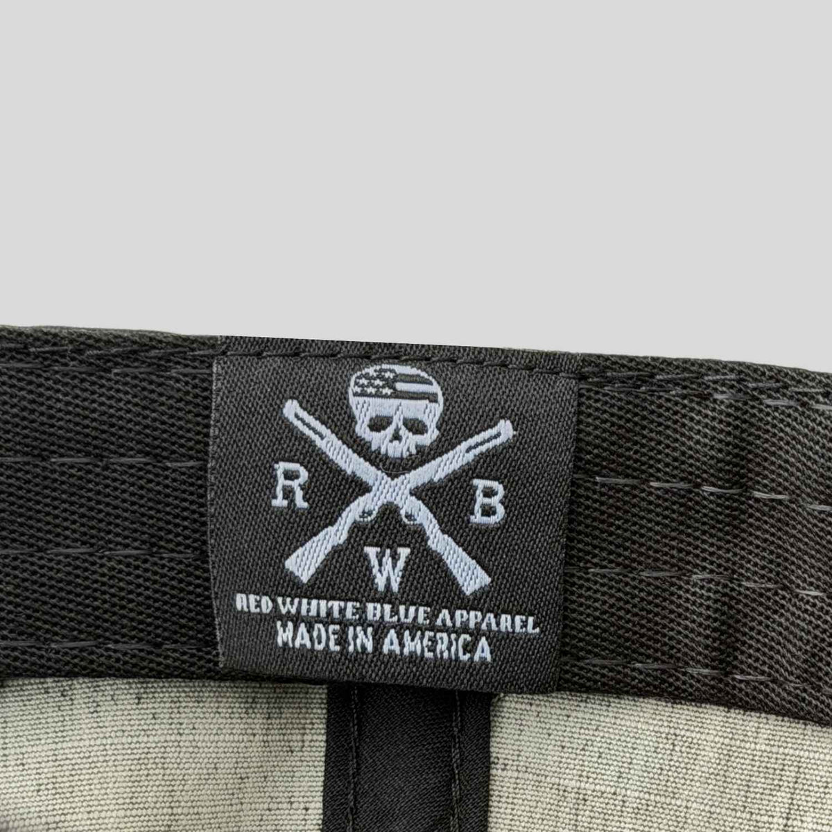 Close-up of a black fabric label sewn onto a black multicam american flag hat, featuring a skull and crossed rifles logo with the text 'RED WHITE BLUE APPAREL MADE IN AMERICA'.