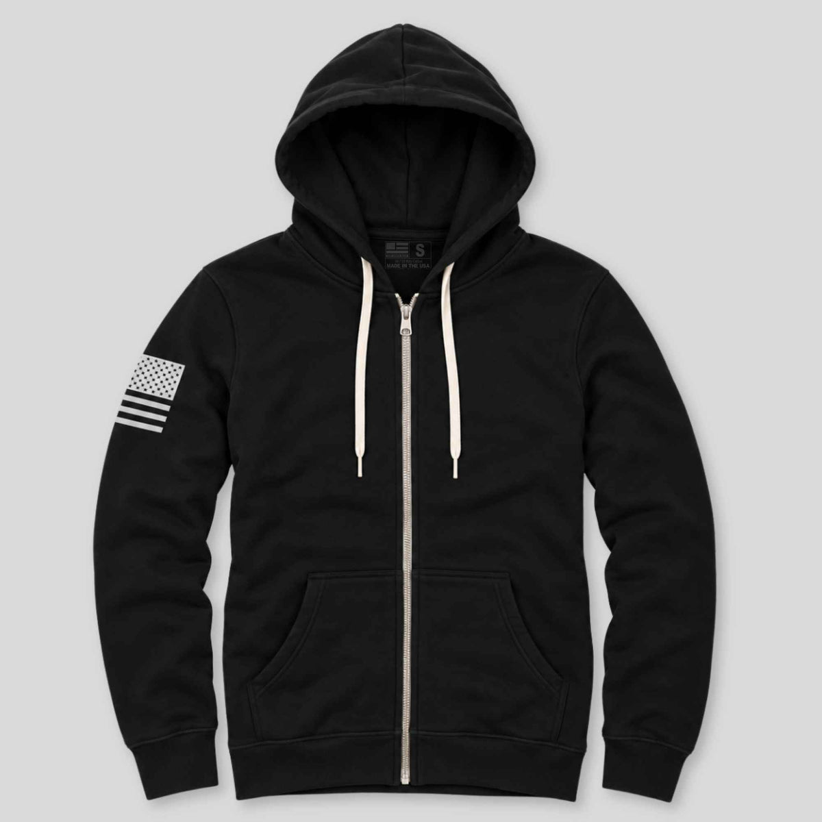 A black zip-up hoodie with cream-colored drawstrings and a white-and-black American flag graphic on the right sleeve, displayed against a solid light gray background.